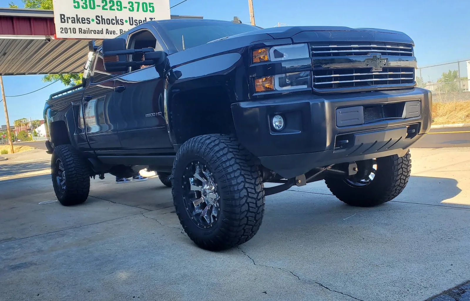 Lifted black Chevy Silverado 2500 HD on chrome wheels and mud-terrain tires under the shop sign